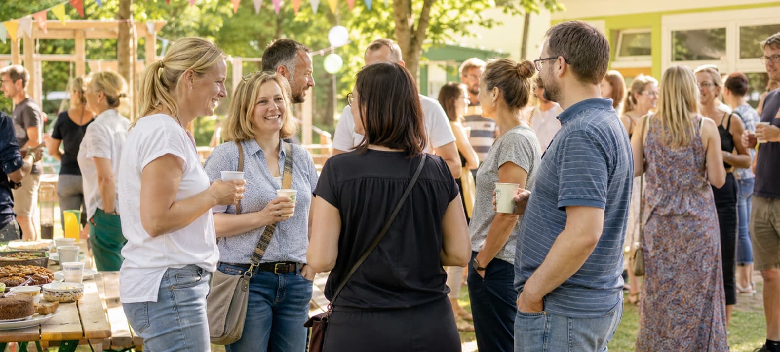 Eltern stehen in Gruppen mit Kaffeebechern beim Sommerfest des Fördervereins auf dem Schulhof, dazwischen Wimpelketten und ein Kuchenbuffet